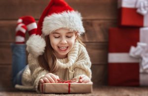 Merry Christmas and Happy Holidays concept. Cute little girl with christmas presents on wooden background. Portrait of beautiful child in Santa hat with gifts.